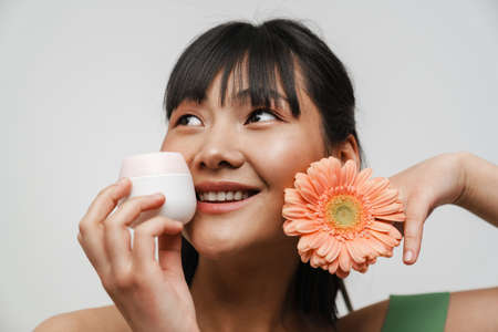 Young asian woman posing with face cream and gerbera flower isolated over white background