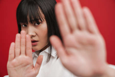 Young asian brunette woman showing stop gesture at camera isolated over red background