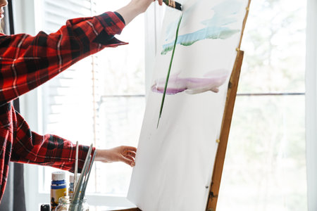 Young white woman in casual wear painting on a canvas indoors near window, holding brush, close up