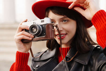Close up of a smiling young stylish brunette white woman wearing hat and leather jacket with short hair standing on a street holding cameraの写真素材