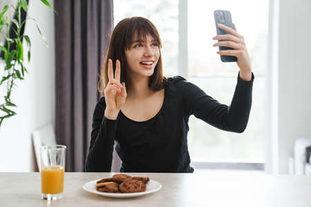 Happy young white woman looking at mobile phone while sitting at the kitchen table at home taking a selfieの写真素材