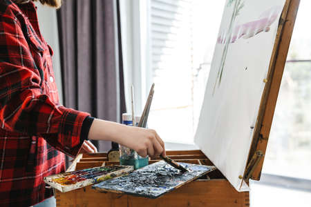 Young white woman in casual wear painting on a canvas indoors near window, close up