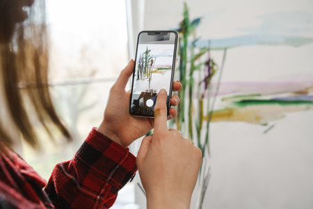 Young white woman painter holding paintbrush standing at the canvas indoors taking picture of canvas close upの写真素材