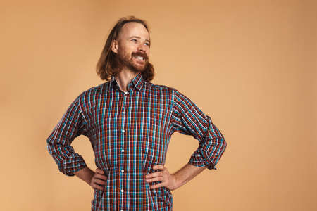 Bearded ginger man wearing plaid shirt smiling and looking aside isolated over beige background