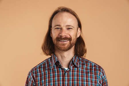 Bearded ginger man wearing plaid shirt smiling and looking at camera isolated over beige background
