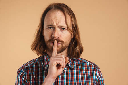 Bearded ginger man frown and showing silence gesture isolated over beige background