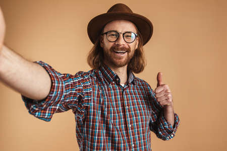 Smiling ginger man in hat showing thumb up and taking selfie photo isolated over beige background