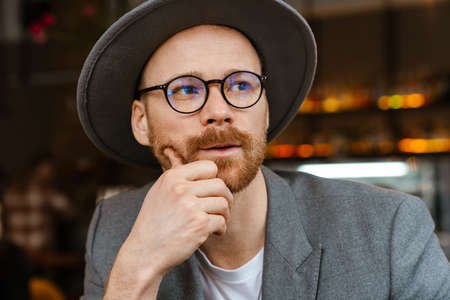 Pensive mid aged fashionable hipster man wearing glasses and hat sitting at the cafe table, looking...