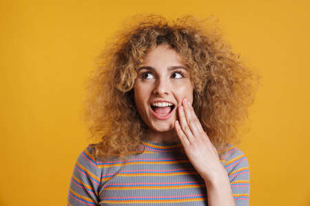Happy smiling young blonde woman with fizzy hair in casual clothes standing over yellow wall background, looking asideの写真素材