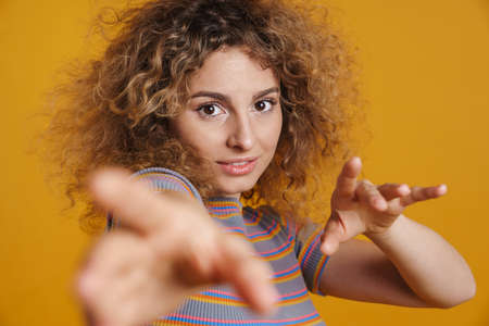 Smiling young casual woman with fizzy hairstyle standing over yellow wall background, holding hands outsretch towards cameraの写真素材