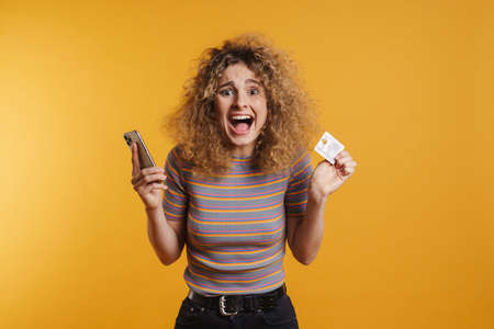 Happy young woman holding mobile phone and credit card celebrating success over yellow wall...