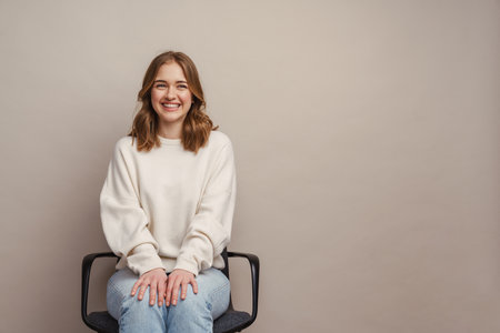 Young white woman smiling while sitting on chair isolated over grey backgroundの写真素材