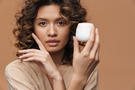 Young curly hispanic woman posing with facial cream isolated over beige backgroundの写真素材
