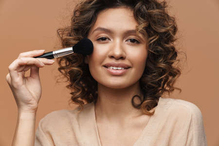 Young curly hispanic woman smiling while using powder brush isolated over beige background