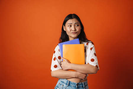 Pensive young asian woman student standing holding textbooks over red wall background lookng asideの写真素材