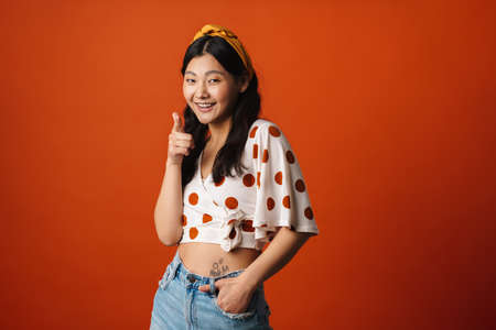Smiling young asian woman in summer clothes standing over orange background