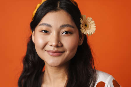 Smiling young asian woman in summer clothes amd floral accessories standing over orange backgroundの写真素材