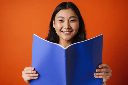 Happy young brunette asian woman holding copybook standing over orange backgroundの写真素材