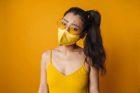 Frowning young asian casual woman wearing protective medical mask standing over yellow background looking at cameraの写真素材
