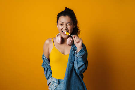 Happy young asian woman with a lollipop wearing summer clothes over orange background, winkingの写真素材