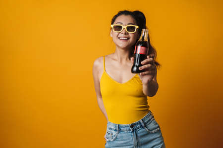 Happy young asian woman wearing summer clothes over orange background, holding bottle with a fizzy drinkの写真素材
