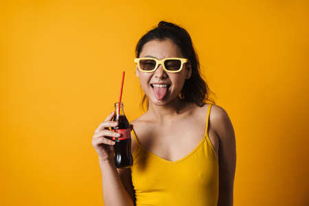 Happy young asian woman wearing summer clothes over orange background, holding bottle with a fizzy drinkの写真素材