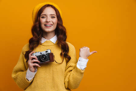 Happy young woman with long red hair in sweater and beret standing over yellow wall background, holding photo camera, pointing finger asideの写真素材