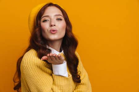 Happy young woman with long red hair in sweater and beret standing over yellow wall background, sending love kissの写真素材