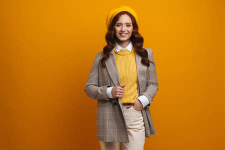 Happy young woman with long red hair in coat and beret standing over yellow wall background looking...