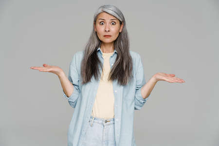 Shocked white-haired woman looking at camera and holding copyspace isolated over gray wall