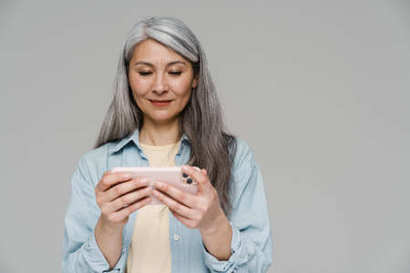 Mature white-haired woman playing online game on mobile phone isolated over gray wallの写真素材