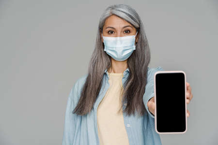 Mature white-haired woman in face mask showing mobile phone isolated over gray wallの写真素材