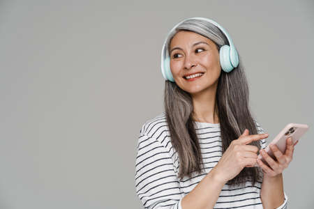 Smiling white-haired woman listening music with cellphone and headphones isolated over gray wall