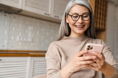 Mature grey woman smiling while using cellphone in kitchen at homeの写真素材