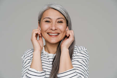 Mature white-haired woman smiling while listening music with earphones isolated over gray wall