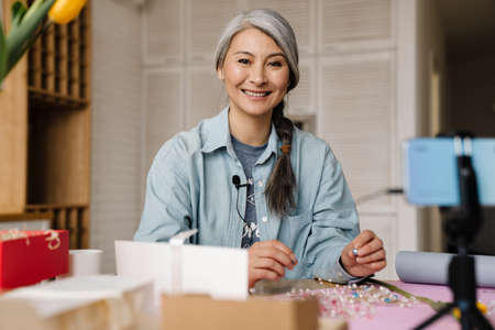 Smiling grey woman showing costume jewelry while taking selfie footage on cellphone at homeの写真素材