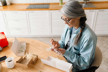 Mature grey woman in eyeglasses wrapping present at homeの写真素材