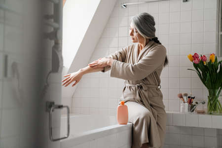Mature grey woman applying cream while sitting in bathroom at home