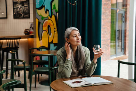 Mature grey woman reading book while drinking water in cafe indoorsの写真素材