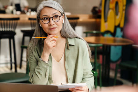 Mature grey woman in earphone working with laptop and papers at cafe indoorsの写真素材