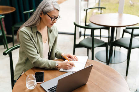 Mature grey woman writing down notes while working with laptop in cafe indoorsの写真素材