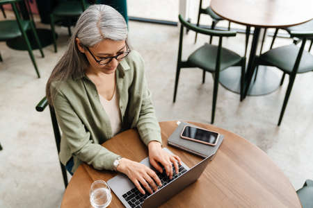 Mature grey woman working with laptop while sitting in cafe indoorsの写真素材