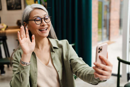 Mature smiling woman gesturing while using earphone and mobile phone in cafe indoorsの写真素材
