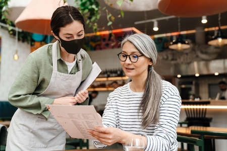 Young waitress woman wearing face mask showing menu to her client in cafe indoorsの写真素材