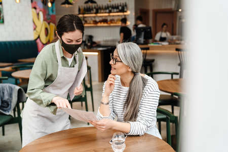 Young waitress woman wearing face mask showing menu to her client in cafe indoors