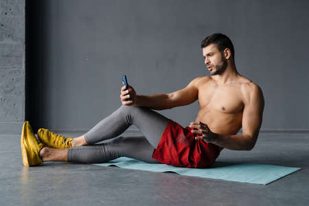 Young shirtless sportsman taking selfie on mobile phone while working out indoors