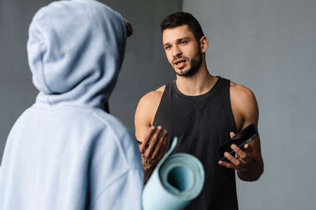 Young fitness coach using cellphone and talking with sportswoman during class indoorsの写真素材