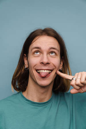 Young white man showing his tongue and pointing finger at his face isolated over blue wallの写真素材