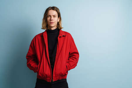 Portrait of a young blonde man in jacket standing over blue background posingの写真素材