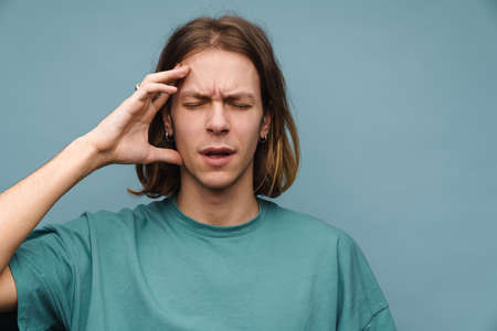 Young white man with headache frowning while holding his head isolated over blue wall